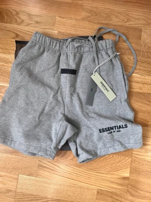 Grå mjukisbyxor från Fear of God - Snygga grå mjukisbyxor från Fear of God Essentials med elastisk midja och dragsko. Byxorna har en avslappnad passform och är tillverkade i mjukt bomullsmaterial. Perfekta för en chill och trendig look.