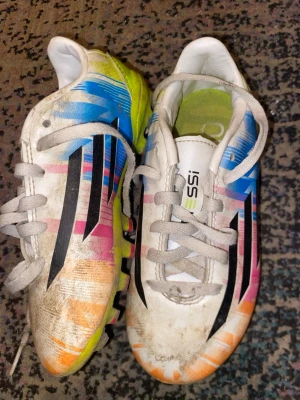 Färgglada Adidas fotbollsskor - Säljer ett par Adidas fotbollsskor med vit bas och färgglada detaljer i blått, rosa, orange och svart. Skorna har klassiska svarta Adidas-ränder på sidorna och snörning framtill. Yttersulan är delvis gul och ovandelen är i syntetmaterial. Perfekta för dig som vill sticka ut på planen!