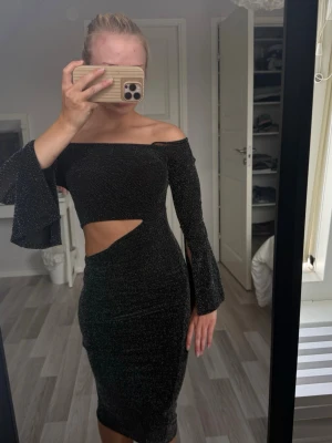 Svart glittrig offshoulder klänning - Säljer en svart glittrig klänning med offshoulder-design och vida ärmar. Klänningen har en snygg cut-out detalj i midjan och sitter tight för en riktigt snygg siluett. Perfekt för dig som vill sticka ut på festen!