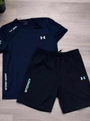 Under Armour träningsset marinblå - Snyggt träningsset från Under Armour i marinblått. Setet består av en t-shirt med rund hals och matchande shorts med dragsko i midjan. Båda plaggen har logga och textdetaljer i vitt och ljusblått. Perfekt för gymmet eller löprundan.