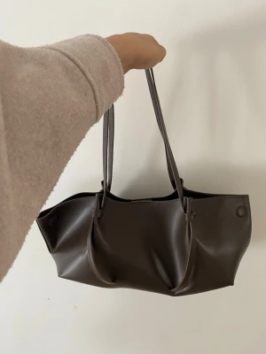 Mörkbrun handväska från Gina Tricot - Stilren mörkbrun handväska från Gina Tricot i slätt konstläder. Väskan har två långa axelband och en mjuk, rymlig form som passar perfekt till alla dina essentials. Enkel och trendig design som funkar till många olika looks.