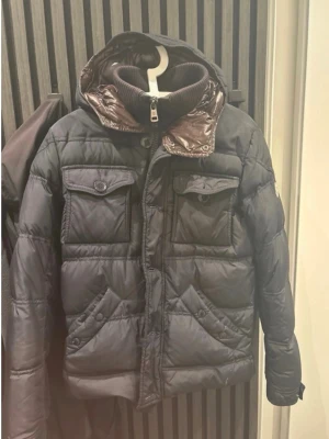 Svart dunjacka från Moncler - Snygg svart dunjacka från Moncler med glansig finish och quiltad design. Jackan har två bröstfickor med lock och knapp, två snedställda fickor framtill samt avtagbar huva och hög krage med dragkedja. Perfekt för kalla vinterdagar.