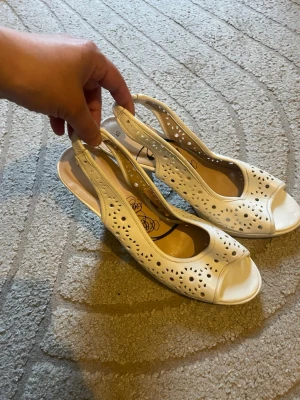 Vita slingback pumps med hålmönster - Snygga vita pumps med öppen tå och slingback-rem. Skorna har ett dekorativt hålmönster i lädret och en stadig klack. Perfekta för dig som vill ha en chic och luftig sko till sommaren. Klacken är medelhög och ger en elegant siluett. Perfekta till midsommar💐💐