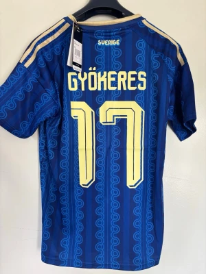 Blå Sverige fotbollströja Adidas - Säljer en blå Sverige fotbollströja från Adidas med Gyökeres och nummer 17 på ryggen. Tröjan har gula detaljer, klassiskt mönster och broderad landslagssköld på bröstet. Tillverkad i lätt och ventilerande material, perfekt för match eller träning.