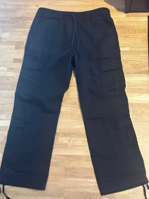 Svarta cargobyxor från GUESS - Säljer ett par svarta cargobyxor från Guess med flera stora fickor på sidorna och dragsko i midjan. Byxorna har avslappnad passform och är perfekta för en streetwear-look. Tillverkade i bomull för skön känsla och slitstyrka.