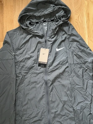 Grå vindjacka med huva från Nike - Snygg grå vindjacka från Nike med huva och dragkedja framtill. Jackan har Nike-logga på bröstet och är tillverkad i lätt, vindtätt material. Perfekt för löpning eller när du vill ha en sportig look. Justerbar huva och diskreta detaljer.