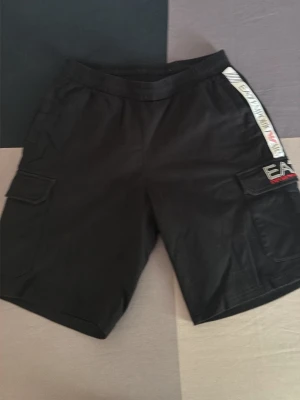 Svarta cargo shorts från EA7 Emporio Armani - Säljer ett par svarta cargo shorts från EA7 Emporio Armani med vita och röda detaljer på sidan. Shortsen har flera fickor, elastisk midja och en relaxed passform. Perfekta för en chill och sportig stil.