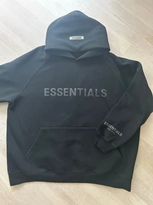 Svart Essentials hoodie med tryck - Säljer en svart hoodie från Essentials med stor logga på bröstet och mindre tryck på armen i reflex. Hoodien har en avslappnad passform och är gjord i mjukt bomullsmaterial. Perfekt för en chill och stilren look.