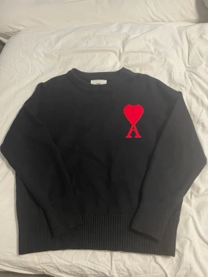 Svart stickad tröja från Ami Paris - Snygg svart stickad tröja från Ami Paris med röd hjärtlogga och bokstaven A på bröstet. Tröjan har rund halsringning, långa ärmar och breda ribbade muddar vid ärmslut och nederkant. Perfekt för dig som gillar stilrena plagg med en twist.