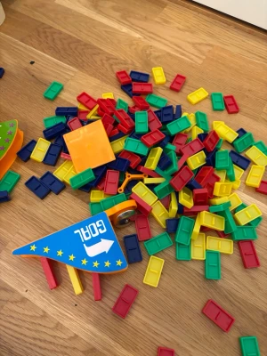 Färgglatt byggspel med plastbitar - Säljer ett byggspel med många rektangulära plastbitar i färgerna röd, gul, grön och blå. Spelet har även två flaggor med texten START och GOAL samt en orange låda för förvaring. Perfekt för kreativ lek och att bygga banor.