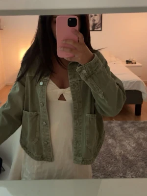 Olivgrön jeansjacka med fickor - Snygg olivgrön jeansjacka med två stora framfickor och klassisk knappstängning. Jackan har en avslappnad passform och raka ärmar, perfekt för en trendig och chill look. Passar till det mesta och ger en cool vibe till outfiten.