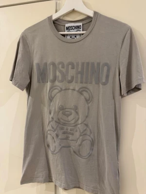 Grå Moschino t-shirt med björntryck - Säljer en grå t-shirt från Moschino med stort ton-i-ton tryck av björn och logga på bröstet. Klassisk rund hals och korta ärmar. Tillverkad i mjuk bomull som känns skön mot huden. Perfekt för dig som gillar streetstyle och statement-plagg.