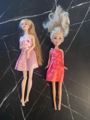 Två dockor med klänningar - Säljer två dockor i plast, båda med blonda hår och klänningar. Ena dockan har långt hår och bär en rosa klänning med hjärtan, den andra har lockigt hår och en enkel rosa klänning. Perfekt för lek eller samling.