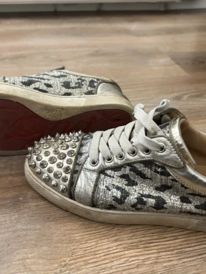 Christian Louboutin leopardmönstrade sneakers - Unika sneakers från Christian Louboutin med silverfärgat leopardmönster, metallicdetaljer och nitar på tån. Skorna har vita snören och den klassiska röda sulan. Perfekta för dig som vill sticka ut med en edgy och lyxig stil.