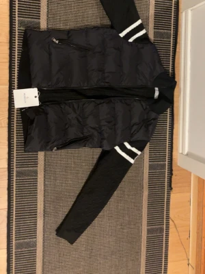 Svart cardiganjacka från Moncler - Svart cardiganjacka från Moncler med vadderad front i nylon och stickade ärmar. Ärmarna har två vita ränder och Moncler-logga på vänster arm. Jackan har dragkedja framtill och en sportig, modern look. Perfekt för lager-på-lager-stil.