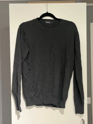 Mörkgrå stickad tröja från Dressmann - Mörkgrå stickad tröja från Dressmann med rund halsringning och långa ärmar. Tröjan har en enkel och stilren design med ribbade muddar vid ärmslut och nederkant. Perfekt basplagg för lager på lager och passar till det mesta. Kan gå ner i pris.