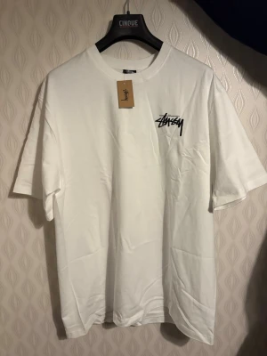 Stüssy t-shirt XL - Säljer en vit t-shirt från Stüssy med svart logga på bröstet och stort tryck på ryggen med rosa tärningar och logga. Klassisk passform och rund hals. Perfekt för dig som gillar streetwear och vill sticka ut med en unik design.