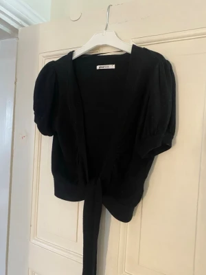 Svart kort kofta från Gina Tricot - Kort svart kofta från Gina Tricot med puffärmar och knytband framtill. Koftan har en djup V-ringning och är gjord i mjukt stickat material. Perfekt att slänga över en topp för en trendig look, storlek S💗