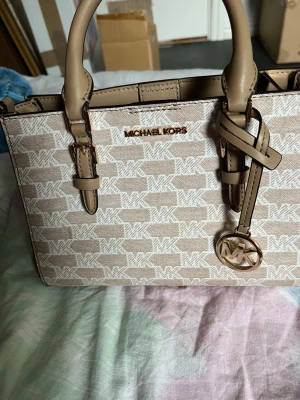 Beige handväska från Michael Kors - Snygg beige handväska från Michael Kors med MK-logga i vitt över hela väskan. Väskan har två handtag, guldfärgade detaljer och en rund MK-berlock. Insidan är ljus med flera fack och dragkedja. Perfekt för dig som gillar stilrena accessoarer.