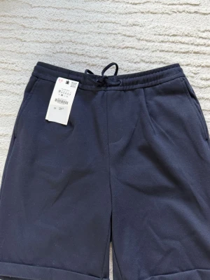 Mörkblå shorts med dragsko - Snygga mörkblå shorts med dragsko i midjan och uppvikta benslut. Shortsen har två sidofickor och är tillverkade i ett mjukt, vävt material som ger en avslappnad look. Perfekta för sommardagar och enkel att matcha med t-shirt eller hoodie.