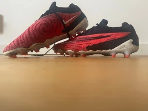 Nike röda fotbollsskor med svart mönster - Säljer ett par Nike fotbollsskor i rött med svart mönster och vita detaljer. Skorna har en låg profil, svart snörning och en sula med dobbar för bra grepp på planen. Swoosh-loggan syns tydligt på sidan och hälen är vit. Perfekta för dig som vill sticka ut på planen. AG Dubbar ( för konstgräs) Elit.