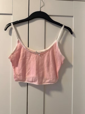 Ljusrosa ribbad linnetopp med rosett - Supersöt ljusrosa linnetopp med tunna vita axelband och en liten rosett framtill. Toppen är ribbad och croppad, perfekt för varma dagar eller som layering-piece. Materialet känns mjukt och lätt mot huden.