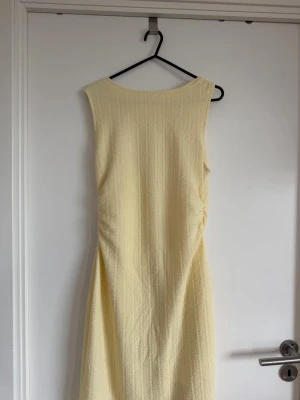 Klänning - Säljer en elegant ljusgul långklänning i ett strukturerat tyg. Klänningen är ärmlös med rund halsringning och har en enkel, stilren design som ger en fräsch och somrig känsla.