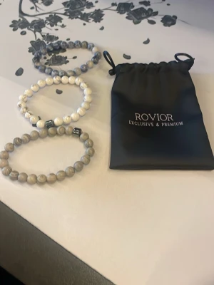 Pärlarmband från Rovior - Tre stilrena armband från Rovior med runda pärlor i olika nyanser av vitt, grått och beige. Armbanden har elastisk tråd och små metalliska detaljer. Kommer med en svart tygpåse med Rovior-logga. Perfekt accessoar för att lyfta din outfit.