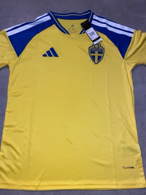 Sverige matchtröja 2026 gul Adidas herr storlek large - Gul matchtröja från Adidas med blå detaljer och Sveriges landslagsemblem på bröstet. Tröjan har blåa axelpartier, vita ränder på axlarna och är tillverkad i lätt, ventilerande ClimaCool-material. Perfekt för fotboll eller träning.
