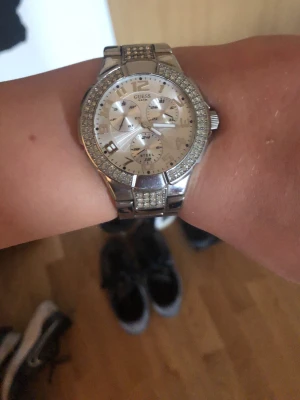 Silverfärgad klocka från Guess - Stilren klocka från Guess i silverfärg med glittrande stenar runt urtavlan och länkarmband i metall. Urtavlan har flera små visare och tydliga siffror, perfekt för dig som gillar en lyxig och modern look.