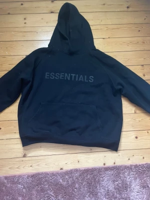 Svart Essentials hoodie - Säljer en svart hoodie från Essentials med stor huva och magficka. Hoodien har trycket 'ESSENTIALS' på bröstet i svart ton-i-ton och är tillverkad i ett mjukt och tjockt material som känns riktigt skönt. Perfekt för dig som gillar streetwear och enkel stil.
