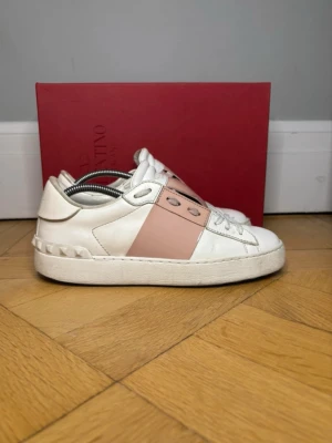 Valentino vita sneakers med rosa detalj - Snygga vita sneakers från Valentino med bred rosa rem över mitten och coola nitar på hälen. Skorna har klassisk låg profil, vita snören och är tillverkade i läder. Perfekta för dig som vill ha en stilren men ändå unik look.