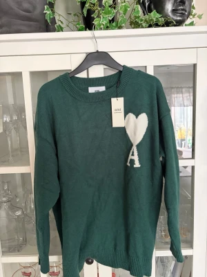 Grön stickad tröja från Ami Paris - Mörkgrön stickad tröja från Ami Paris med rund halsringning och ribbade muddar. På bröstet finns ett vitt hjärta och bokstaven A i kontrastfärg. Tröjan har en avslappnad passform och är tillverkad i mjukt material.