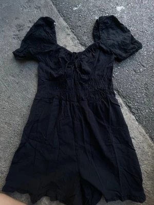 Svart jumpsuit med puffärm - Supersnygg svart jumpsuit med korta ben och puffiga, broderade ärmar. Smock i midjan och söt detalj med knytning vid bysten. Tillverkad i ett lätt tyg som känns luftigt och skönt. Perfekt för dig som gillar trendiga detaljer och en avslappnad look.