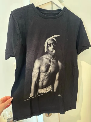 Svart t-shirt med Tupac-tryck - Cool svart t-shirt med stort tryck av Tupac på framsidan. Klassisk rund hals och korta ärmar.