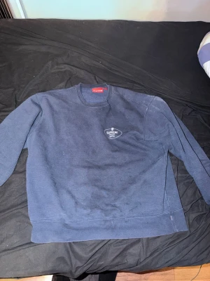 Supreme Crest Crewneck Navy - Supreme sweatshirt i mörkblått med rund halsringning och tryckt logga på bröstet samt stor Supreme New York-print på ryggen. Klassisk passform och mjukt material, perfekt för en avslappnad streetwear-look.