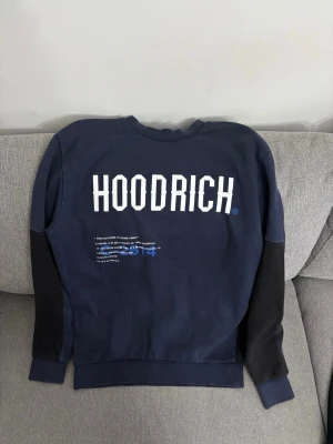 Blå sweatshirt från Hoodrich - Cool blå sweatshirt från Hoodrich med stora vita tryck på ryggen och svarta detaljer på ärmar och nedre delen av tröjan. Tröjan har rund hals och tryck med världskarta och text, perfekt för en streetwear-look.