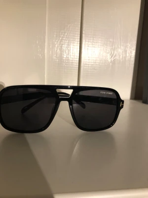 Svarta Tom Ford solglasögon - Snygga svarta solglasögon från Tom Ford med rektangulär form och mörka glas. Klassisk design med gulddetalj vid skalmen och logga på insidan. Perfekta för dig som vill ha en stilren och exklusiv look.