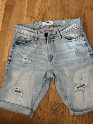 Ljusblå slitna jeansshorts från Fish - Säljer ett par ljusblå jeansshorts från Fish med slitna detaljer och upprullade benslut. Shortsen har fem fickor, knappgylf och klassisk jeanslook. Perfekta för en avslappnad stil i sommar.