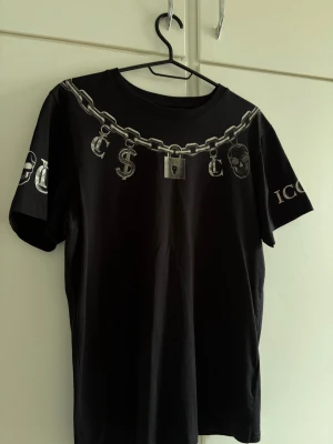 Svart t-shirt med kedjetryck och döskalle - Cool svart t-shirt med tryck av en stor silverfärgad kedja, hänglås, dollartecken och döskalle över bröstet. 