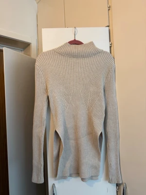 Ribbstickad polotröja från NA-KD - Säljer en beige ribbstickad polotröja med lång ärm och hög krage. Tröjan har en figurnära passform och är tillverkad i ett mjukt, stretchigt material som ger en snygg siluett. Det är slitasar på sidorna som börjar vid höften.