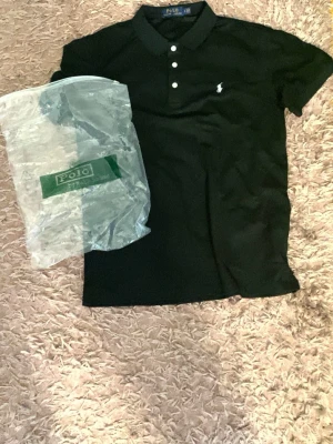 Svart pikétröja från Polo Ralph Lauren - Klassisk svart pikétröja från Polo Ralph Lauren med vit broderad logga på bröstet. Tröjan har krage, tre vita knappar framtill och korta ärmar. Perfekt till både jeans och shorts för en clean look.