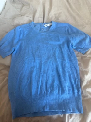 Blå kortärmad topp från Zara - Snygg blå topp från Zara med korta ärmar och rund halsringning. Toppen har ribbade detaljer vid nederkant och ärmslut samt dekorativa knappar i nacken. Perfekt för en stilren och enkel look.