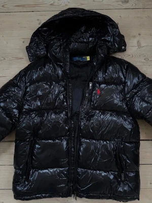 Svart dunjacka från Polo Ralph Lauren - Snygg svart dunjacka från Polo Ralph Lauren med glansig finish och röd broderad logga på bröstet. Jackan har huva, dragkedja framtill och flera praktiska fickor. Perfekt för kalla vinterdagar och riktigt stilren look.