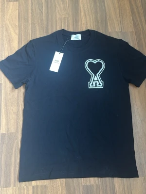 Svart t-shirt från Ami Paris - Snygg svart t-shirt från Ami Paris med det ikoniska hjärtlogotypen broderad på bröstet. Klassisk passform och rund halsringning. Tillverkad i mjuk bomull för maximal komfort. Perfekt för en clean och stilren look.