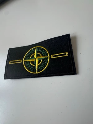 Stone Island badge - Säljer ett svart tygmärke från Stone Island med broderad kompasslogga i gult och grönt. Märket är rektangulärt och passar perfekt att sy eller fästa på jackor, tröjor eller väskor för en cool streetwear-look.