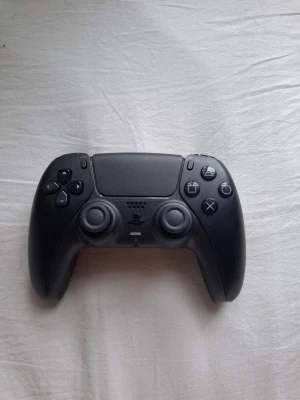 Svart PlayStation 5 handkontroll - Säljer en svart PlayStation 5 DualSense handkontroll från Sony. Den har en modern design med touchpad, två analoga spakar och klassiska PlayStation-knappar. Tillverkad i matt och blank plast för skön känsla i handen. Perfekt för gaming på PS5.