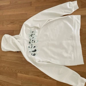 Vit hoodie från Off-White - Säljer en vit hoodie från Off-White med svart logotryck på bröstet och grafiskt motiv på ryggen. Hoodien har svarta snören i huvan och en stor magficka. Materialet är mjukt och bekvämt, perfekt för en avslappnad look.