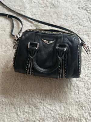 Svart handväska från Zadig & Voltaire - Cool svart handväska från Zadig & Voltaire i äkta läder med silverfärgade detaljer och nitar. Väskan har två handtag, avtagbar axelrem och dekorativa dragkedjor framtill. Perfekt för dig som gillar rockig stil och snygga accessoarer.