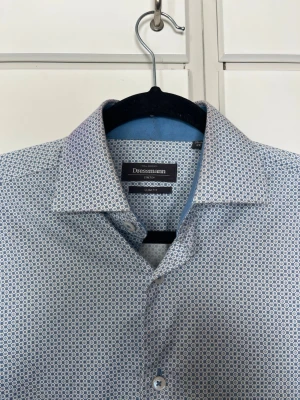 Mönstrad blå skjorta från Dressmann - Snygg långärmad skjorta från Dressmann med diskret blått och vitt mönster. Skjortan har klassisk krage, knappar framtill och är i slim fit-modell. Perfekt för dig som gillar stilrena detaljer och vill ha en skjorta som sticker ut lite extra.
